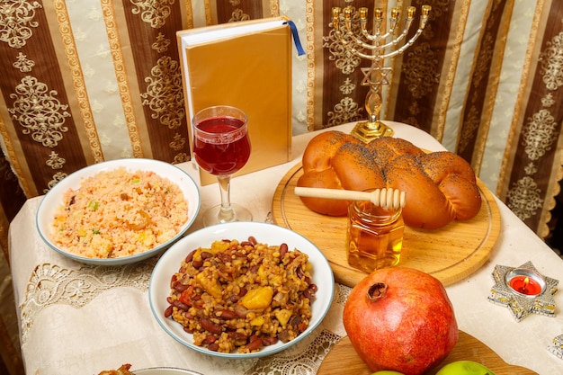 Mesa festiva para una comida en Rosh Hashanah challah miel granada manzanas vino y un chelnt tradicional junto a la menorá y siddur