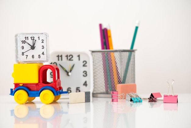 En la mesa frente al reloj y un vaso con lápices hay un carro de juguete con un reloj en la parte superior peinados de papelería y un sacapuntas y molinillos están dispersos a un lado