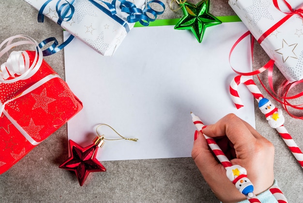 Una mesa gris con una hoja de felicitación, adornos navideños, una taza de chocolate caliente y un bolígrafo en forma de bastón de caramelo. niña escribiendo, mano femenina en la imagen, vista superior