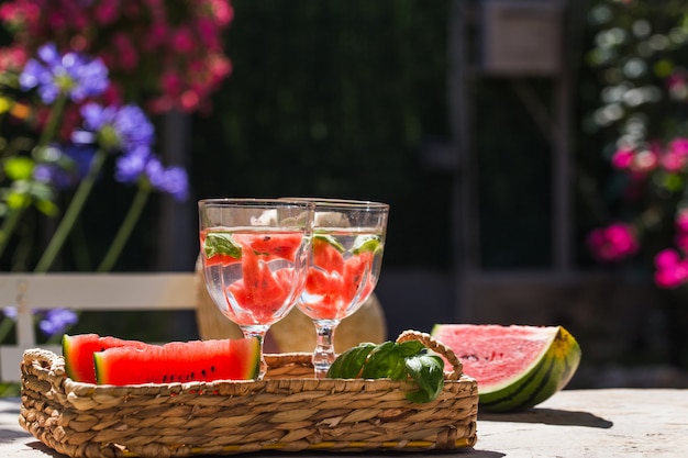 En una mesa en el jardín en una canasta hay dos vasos de limonada y rodajas de sandía