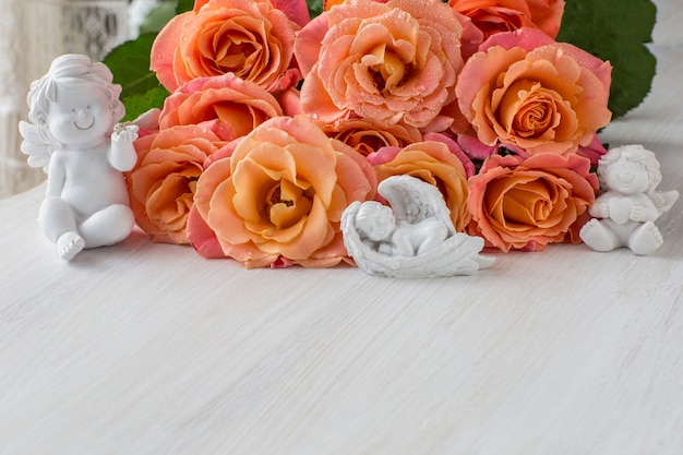 En una mesa de luz, un ramo de rosas con rosas naranjas y tres ángeles blancos.