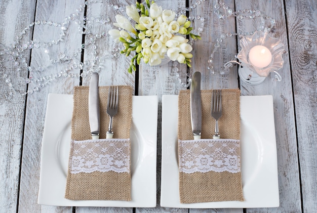 Foto en una mesa de madera blanca, dos platos, velas, tenedores y cuchillos, flores en un jarrón: un fondo festivo (cumpleaños, boda, 8 de marzo, cena romántica)