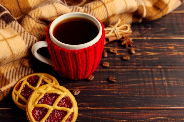 En una mesa de madera cerca de la tela escocesa a cuadros hay una taza de café caliente, con una cubierta tejida y galletas y especias caseras.