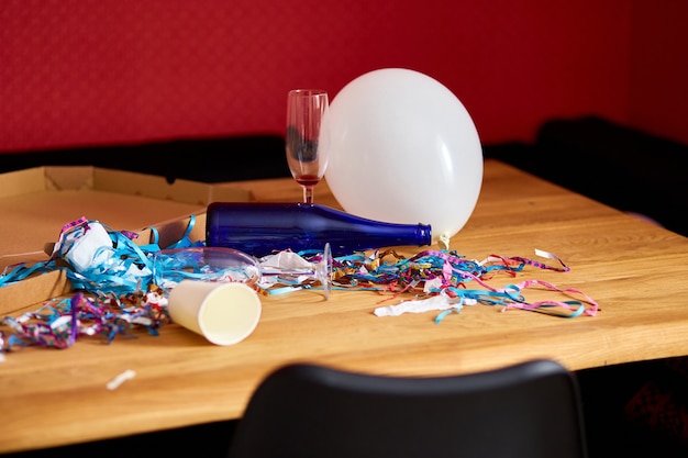 Mesa de madera desordenada con caja de pizza, botellas azules vacías, copa de vino y decoraciones de fiesta en la mañana después de la celebración de la fiesta, después del caos de la fiesta, sobras de la celebración.