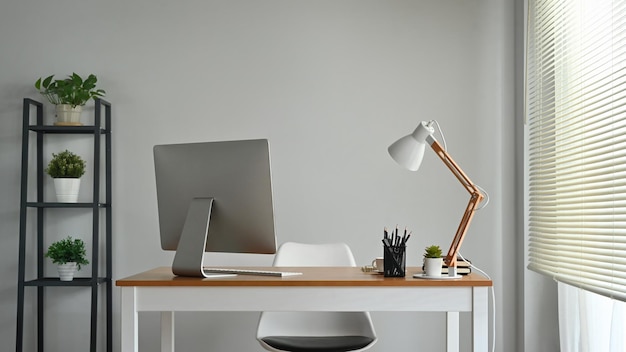 Mesa de madera con monitor de computadora y artículos de papelería y lámpara en el acogedor interior de la oficina decorado con plantas en macetas verdes