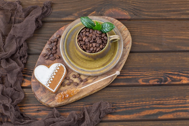 en una mesa de madera una taza con granos de café, un pan de jengibre en forma de corazón y azúcar