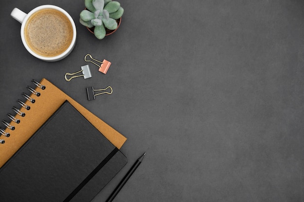 Mesa moderna e minimalista com cadernos, xícara de café, planta suculenta verde sobre fundo escuro texturizado. Escritório ou espaço de trabalho de negócios com espaço de cópia de texto. Lay criativo apartamento.