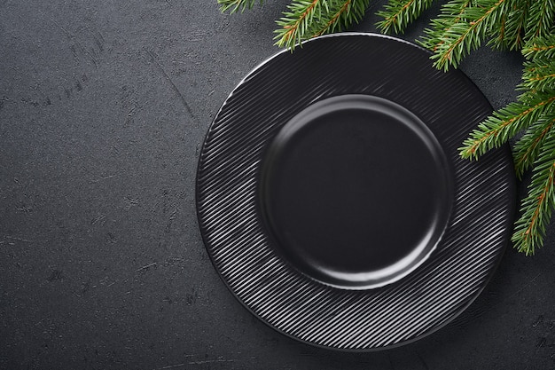 Mesa de navidad servida con plato de cerámica negro vacío sobre fondo oscuro. Decoración navideña. Decoraciones navideñas con ramas de abeto sobre fondo negro oscuro. Diseño de borde. Vista superior.