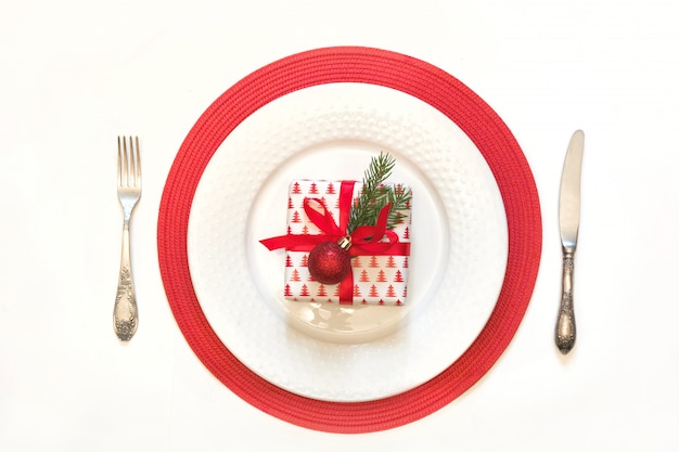 Mesa de Navidad con vajilla blanca, cubiertos y decoraciones rojas sobre fondo blanco.