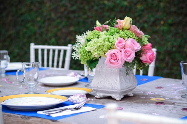 Mesa no jardim com pratos de porcelana azul e amarelo com copos de cristal vazios e um arranjo de flores no centro com rosas cor de rosa e cadeiras brancas há desfoque no fundo