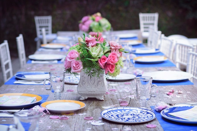 Mesa no jardim com pratos de porcelana azul e amarelo com copos de cristal vazios e um arranjo de flores no centro com rosas cor de rosa e cadeiras brancas há desfoque no fundo