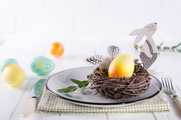 Mesa de Pascua con plato blanco, servilleta textil, huevo de gallina amarillo decorativo en el nido, flores de mimosa, plumas y decoración de Pascua en la mesa blanca