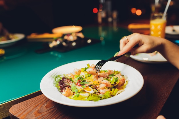 Mesa de restaurante o cafetería con plato de ensaladas y vino. Dos personas hablando sobre fondo. Imagen entonada