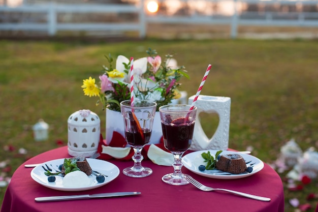 Mesa romántica para dos al aire libre con mantel rojo y cubiertas en sillas con postres vino caliente un ramo de flores velas y pétalos de rosa El concepto de propuesta de matrimonio