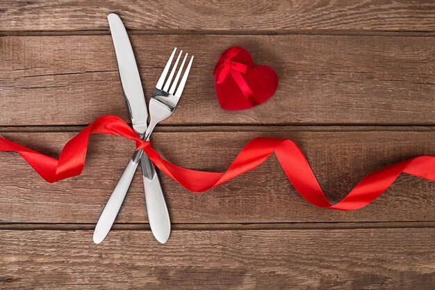 Mesa de San Valentín con cinta roja, cuchillo y tenedor y anillo sobre fondo de roble. Naturaleza muerta