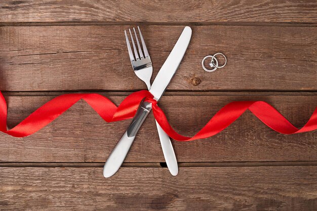 Mesa de San Valentín con cinta roja, cuchillo y tenedor y anillo sobre fondo de roble. Naturaleza muerta