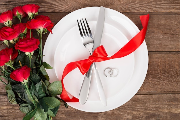 Mesa de San Valentín con cinta roja, rosas, cuchillo y tenedor y anillo sobre fondo de roble. Naturaleza muerta