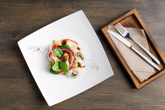 Mesa servida com salada de polvo e legumes na chapa branca, cópia espaço, vista superior. Refeição apetitosa de frutos do mar mediterrâneos frescos e talheres em caixa de madeira. Serviço de restaurante