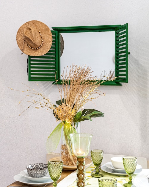 Una mesa servida con platos, vasos y flores decorativas en florero. Comedor con espejo y sombrero de paja.