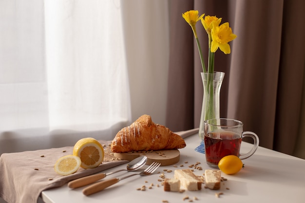 Mesa servindo café da manhã com uma xícara de chá, croissants, limões e narcisos amarelos em um vaso de vidro