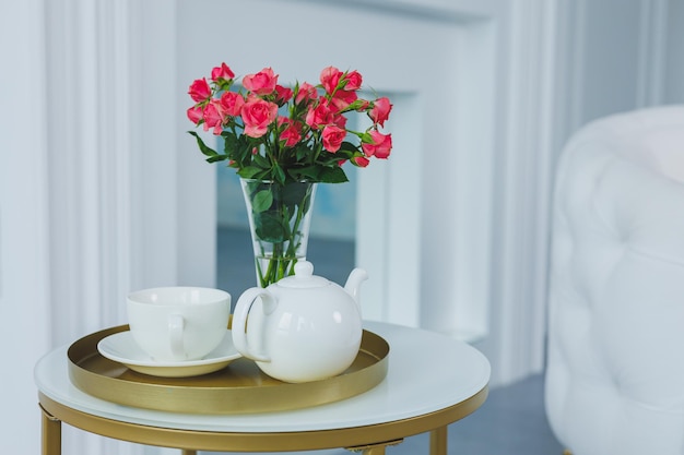 Una mesa con una tetera blanca y una taza blanca sobre la mesa un jarrón con flores frescas