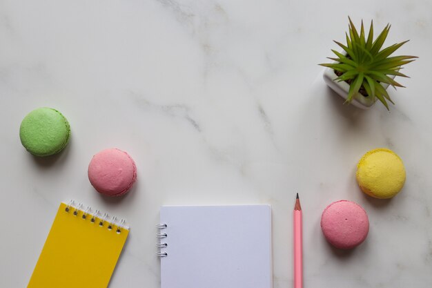 Mesa de trabajo con blocs de notas, lápiz, planta y macarrones sobre fondo de mármol con espacio de copia