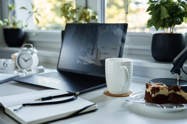 Foto mesa de trabajo de oficina en casa con portátil y café caliente cerca de la ventana en la sala de estar