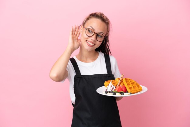 Mesero de restaurante Chica rusa sosteniendo gofres aislados en un fondo rosa escuchando algo poniendo la mano en la oreja