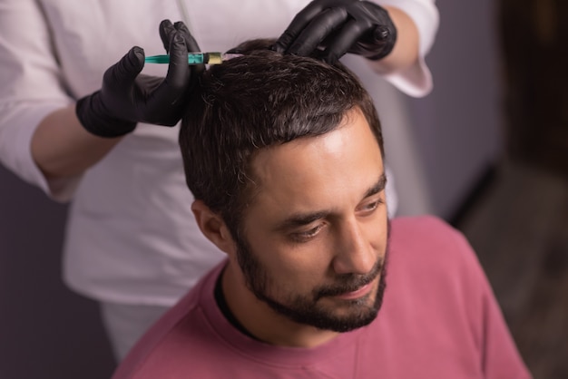 Mesotherapie für Haare. Attraktiver Mann, der Injektionen in seinen Kopf erhält. Mann mit Mesotherapie-Sitzung im Schönheitssalon, Therapeut im Schutzhandschuh mit Spritze,