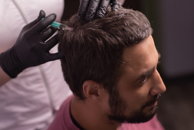 Mesotherapie für Haare. Attraktiver Mann, der Injektionen in seinen Kopf erhält. Mann mit Mesotherapie-Sitzung im Schönheitssalon, Therapeut im Schutzhandschuh mit Spritze,