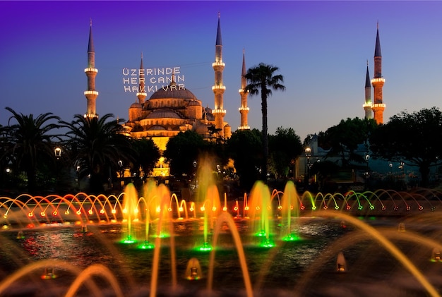 Mesquita azul e fonte em Istambul iluminada à noite, Turquia. A inscrição em uma mesquita é traduzida como "Nele está a alma de todos".