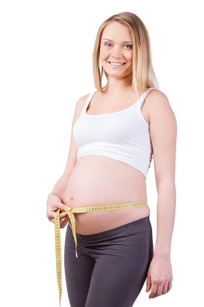 Messen ihres schwangeren Bauches. Schöne schwangere Frau, die ihren schwangeren Bauch misst und lächelt, während sie isoliert auf weiß steht