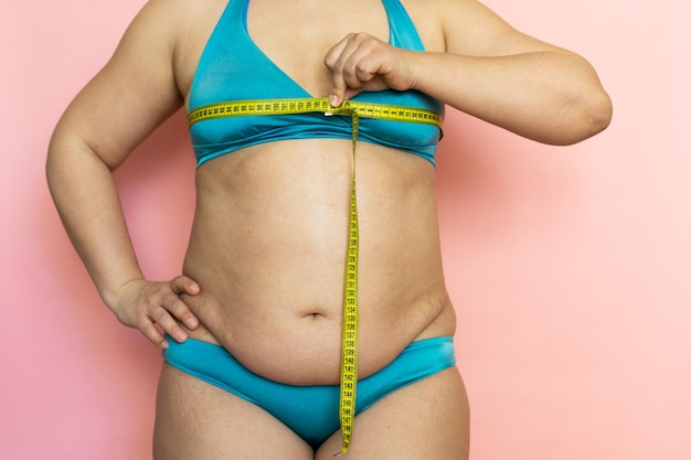 Messen Sie die weibliche Brust mit Klebeband und bestimmen Sie die Anzahl der Nahaufnahmefalten auf dem Bauch Cellulite Nacktes übergewichtiges plus Größenmädchen auf rosa Hintergrund in blauer Unterwäsche Konzept der Diät und Körperbeherrschung