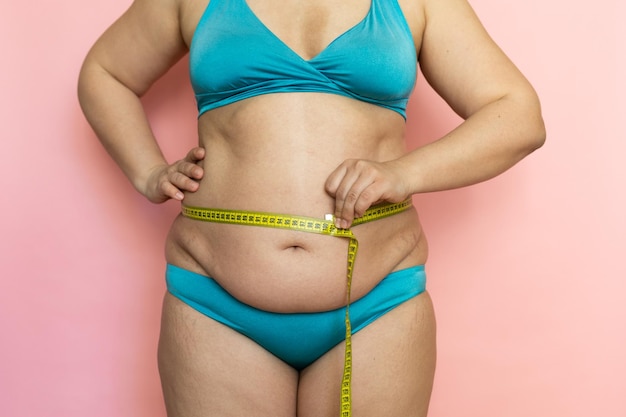 Foto messen sie die weibliche taille mit klebeband nahaufnahmefalten am bauch lockere haut fettleibigkeit und cellulite nacktes übergewichtiges plus größenmädchen auf rosa hintergrund in blauer unterwäsche konzept der diät und körperbeherrschung