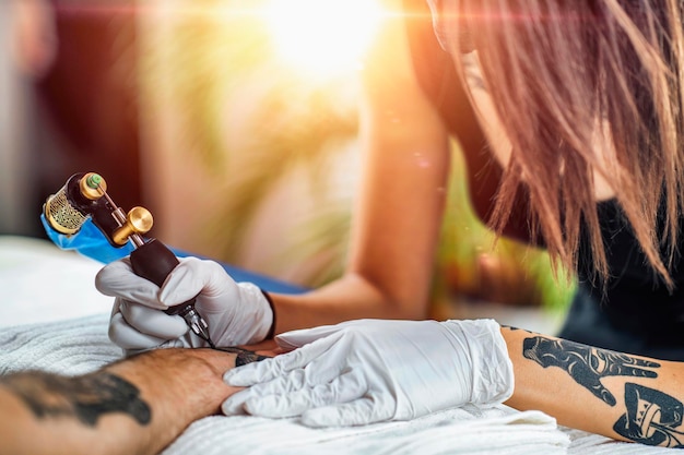 Mestre de tatuagem feminina profissional aplicando tatuagem na mão de jovem com máquina de tatuagem