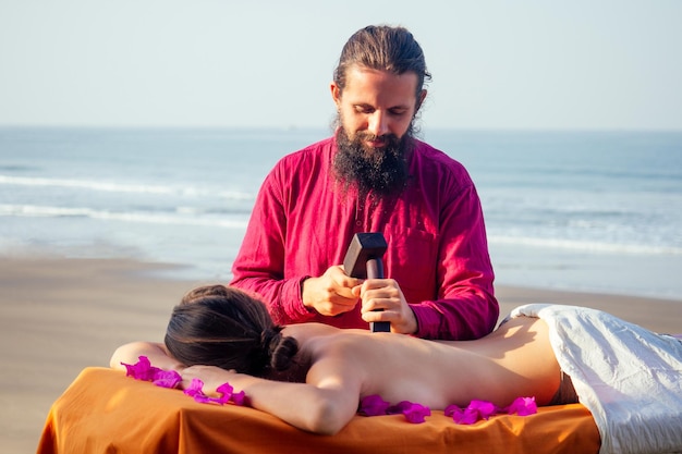 Mestre masculino faz massagem tailandesa Tok-sen spa terapia fisiologia pela praia do oceano do mar Mulher bonita tendo uma massagem relaxante nas costas tailandesa com conjunto para massagem toksen