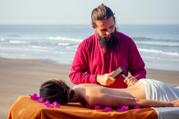 Mestre masculino faz massagem tailandesa Tok-sen spa terapia fisiologia pela praia do oceano do mar Mulher bonita tendo uma massagem relaxante nas costas tailandesa com conjunto para massagem toksen