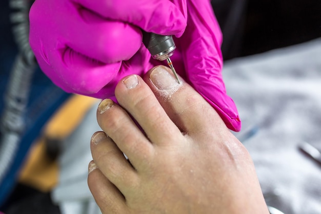 Mestre pedicure em luvas cor de rosa corta as unhas da cutícula e goma-laca no salão de pedicure usando broca Pedicure profissional na clínica de cosmetologia Higiene para os pés no salão de beleza