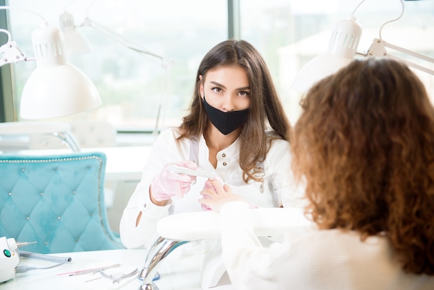 Mestre profissional de manicure em máscara protetora usando ferramenta elétrica de polidor de unhas para procedimento de manicure de tratamento de vidro.