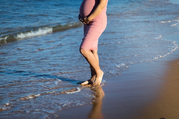 metade do corpo da mulher grávida no vestido rosa segurando a barriga e caminhar na praia