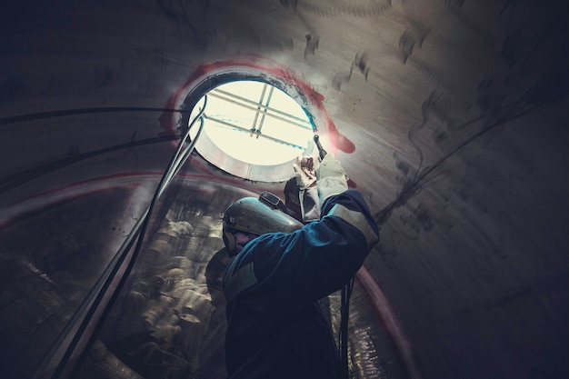 El metal reparado masculino del trabajador del argón del arco de soldadura está soldando chispas del tanque de la construcción industrial parte del pozo del aceite inoxidable dentro de los espacios confinados hay un vigilante afuera.