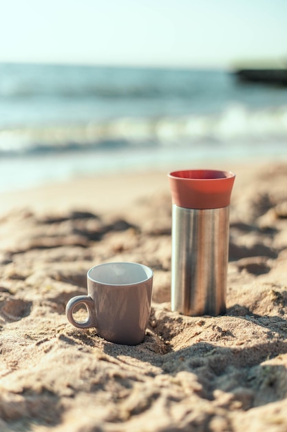 Metall-Thermoskanne oder Thermoskanne mit Kaffee- oder Teegetränk an der einsamen Sandküste des Ozeans oder Meeres