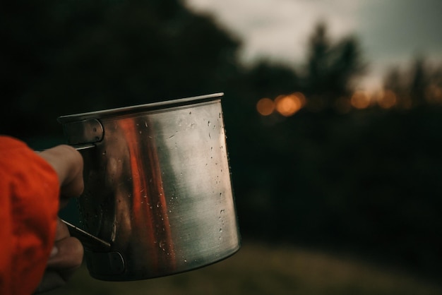 Metallbecher mit Tee in der Hand eines Touristen im Zeltlager in Bergen unter weißen Wolken