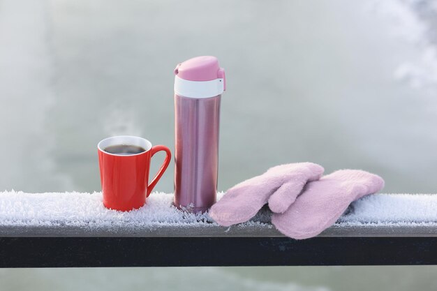 Metallrosa Thermoskanne mit gestrickten Fäustlingen und einer roten Tasse heißen Tees auf dem Hintergrund der Winternatur Selektiver Fokus