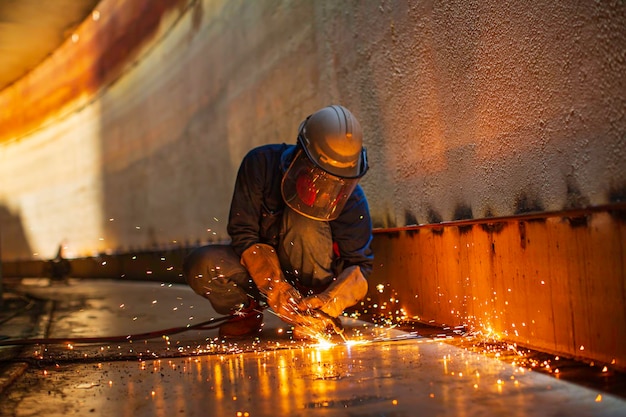 Metallschneidender Funke des männlichen Arbeiters auf der Stahlplatte des Tankbodens mit Blitz des Schneidlichts aus nächster Nähe, Schutzhandschuhe und Maske tragen