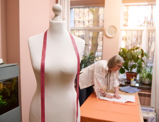 Meter und Schaufensterpuppe im Modedesign-Atelier auf verschwommenem Hintergrund einer Schneiderin, die Schnittmuster auf Gewebe überträgt, während sie neue Kleidung kreiert
