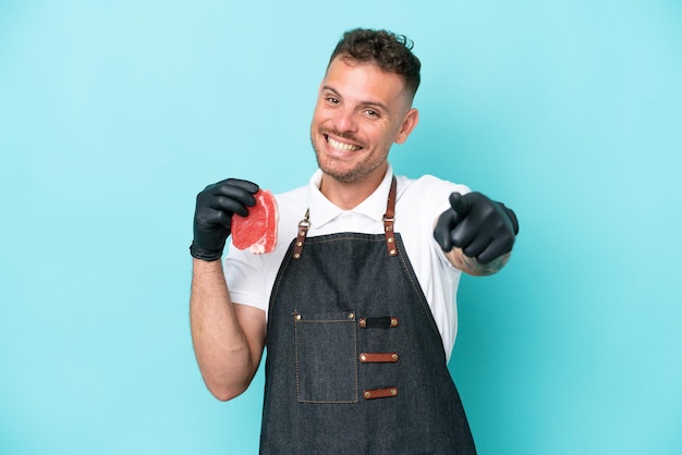 Metzger, kaukasischer Mann, der eine Schürze trägt und frisch geschnittenes Fleisch serviert, isoliert auf blauem Hintergrund, der mit glücklichem Ausdruck nach vorne zeigt