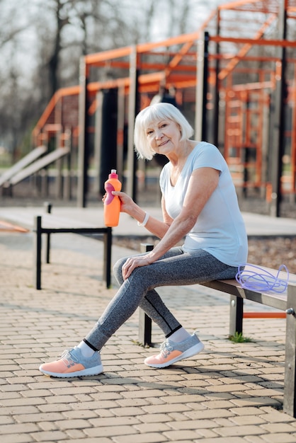 Meu descanso. Mulher feliz e magra segurando uma garrafa de suco e relaxando enquanto se exercita