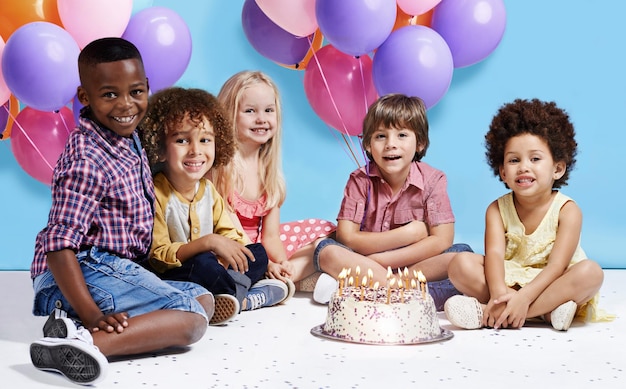 Meu desejo de aniversário é sermos amigos para sempre Foto de um grupo de crianças sentadas ao redor de um bolo de aniversário com um monte de balões ao fundo