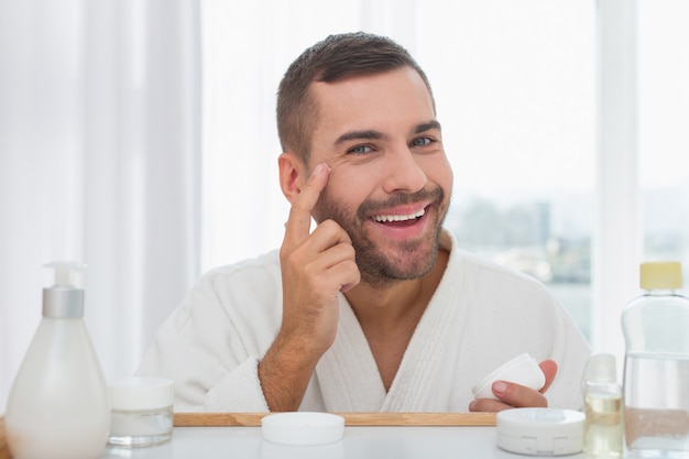 Meu rosto. Homem bonito alegre sorrindo enquanto passa creme no rosto